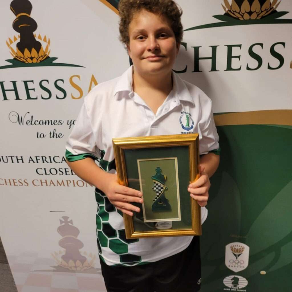 A person wearing a chess-themed shirt holds a framed decoration and stands in front of a ChessSA banner at a South African Chess Championship event, highlighting the rise of online chess classes.