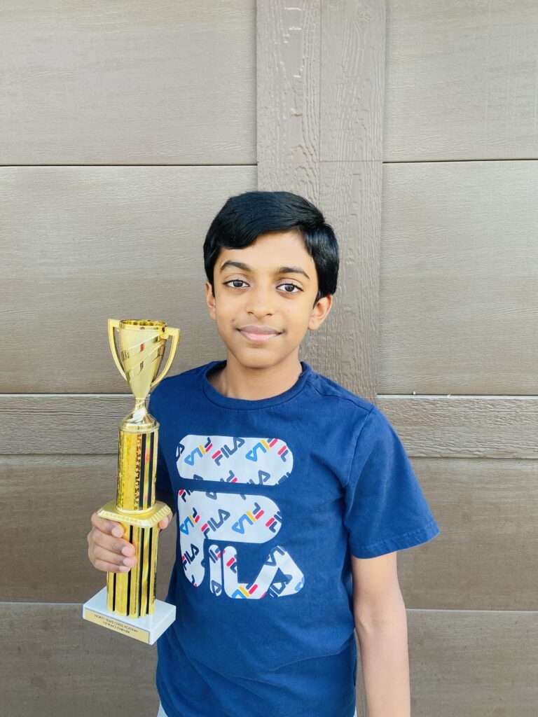 A young boy smiling and holding a large trophy, standing in front of a beige wall. He wears a blue T-shirt with colorful designs, celebrating his victory in online chess lessons.