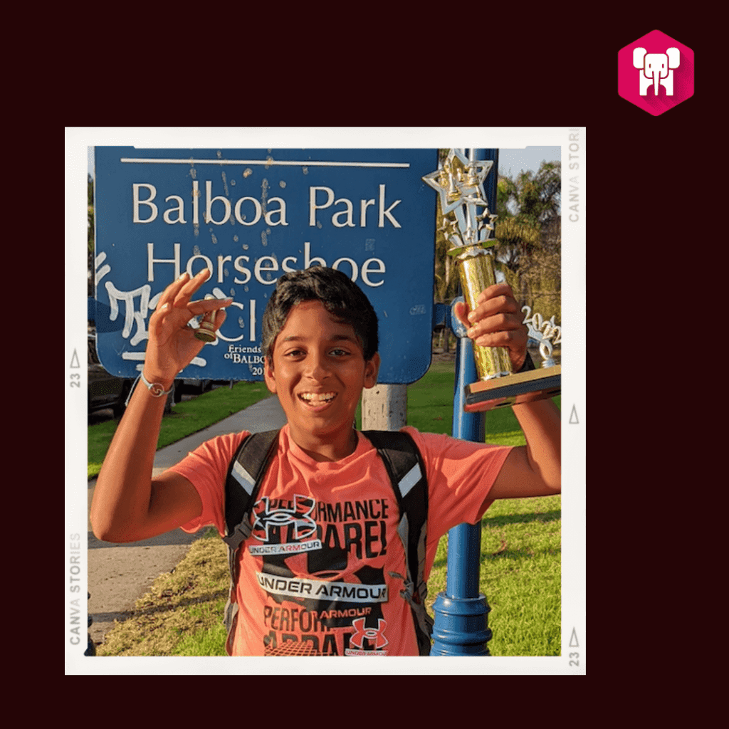 A joyful boy holding a trophy in front of a Balboa Park Horseshoe sign, wearing a bright orange shirt after winning at his chess coaching session.