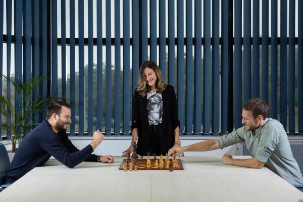 Two men play chess at a table while a woman stands between them, observing and smiling. They are in a bright room with large windows and venetian blinds.