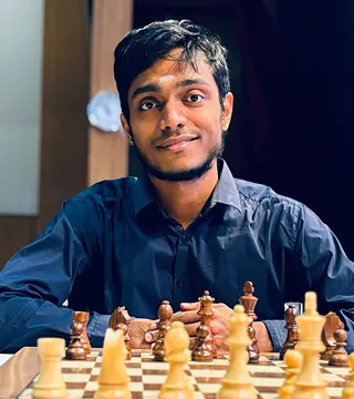 A man in a dark shirt sits at a chessboard, looking at the camera with a slight smile; chess pieces are arranged in front of him.