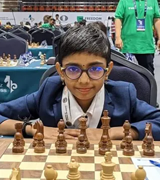 A young person wearing glasses and a suit jacket sits at a chessboard in a tournament hall, looking at the camera.