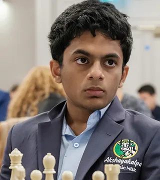 A young person in a blazer sits at a chessboard, looking focused. The blazer has an "Akshayakalpa Organic Milk" logo on it. Other people are visible in the background.