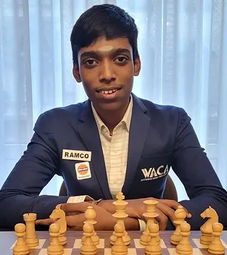 A young man in a blue suit sits at a chessboard with chess pieces arranged, looking at the camera.
