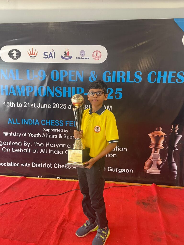 A young boy in a yellow shirt holds a large trophy, standing in front of a banner for the National U-9 Open & Girls Chess Championship 2025.