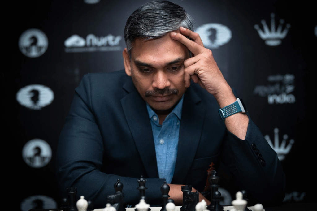 A man in a suit concentrates on a chessboard, resting his head on one hand, with chess pieces and a dark, branded background visible.