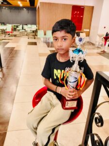A boy sits on a red chair indoors, holding a large trophy. He wears a black t-shirt and light-colored pants, with an expressionless face.