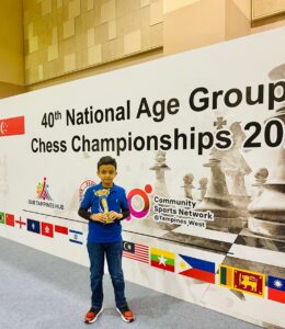 A boy holding a trophy stands in front of a wall displaying "40th National Age Group Chess Championships" and international flags.