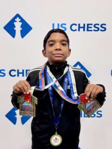 A young boy stands in front of a US Chess backdrop, holding two large medals and wearing a third medal around his neck.
