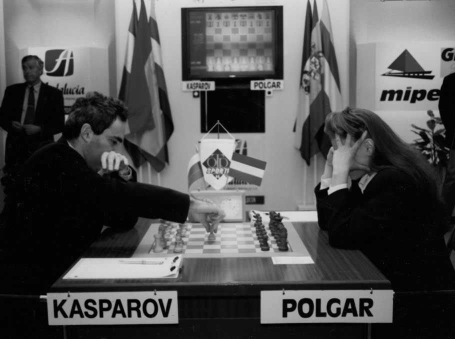 Two chess players, Garry Kasparov and Judit Polgar, play a game at a tournament table with flags and an electronic board displayed in the background.