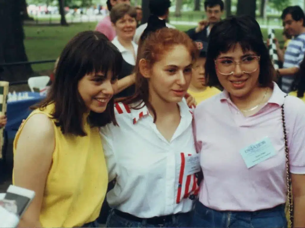 Three young women stand close together and smile for a photo at an outdoor event. Other people and tables are visible in the background.
