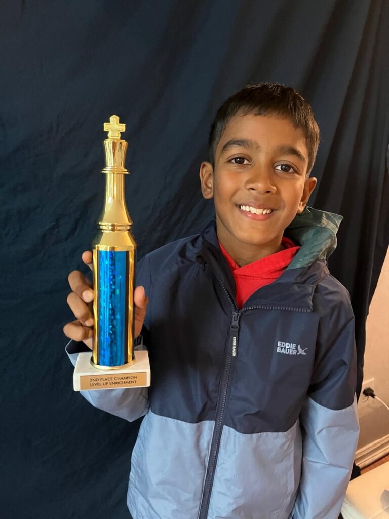 A boy in a jacket smiles and holds a gold and blue trophy with a plaque that reads “2ND PLACE CHAMPION LEVEL 2 CHESS TOURNAMENT.”.