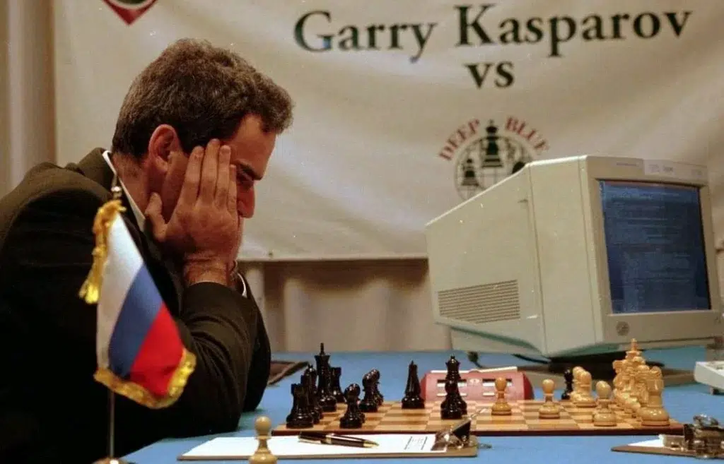 A man plays chess against a computer at a table with a Russian flag; a banner behind reads "Garry Kasparov vs Deep Blue.
