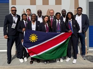A group of eleven people in formal attire pose together outdoors, holding a Namibian flag in front of them.
