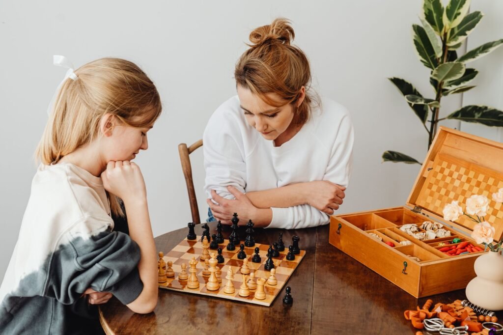 A mother sits alongside her daughter studying a chess board at a home table, illustrating the parent's supportive role in chess development