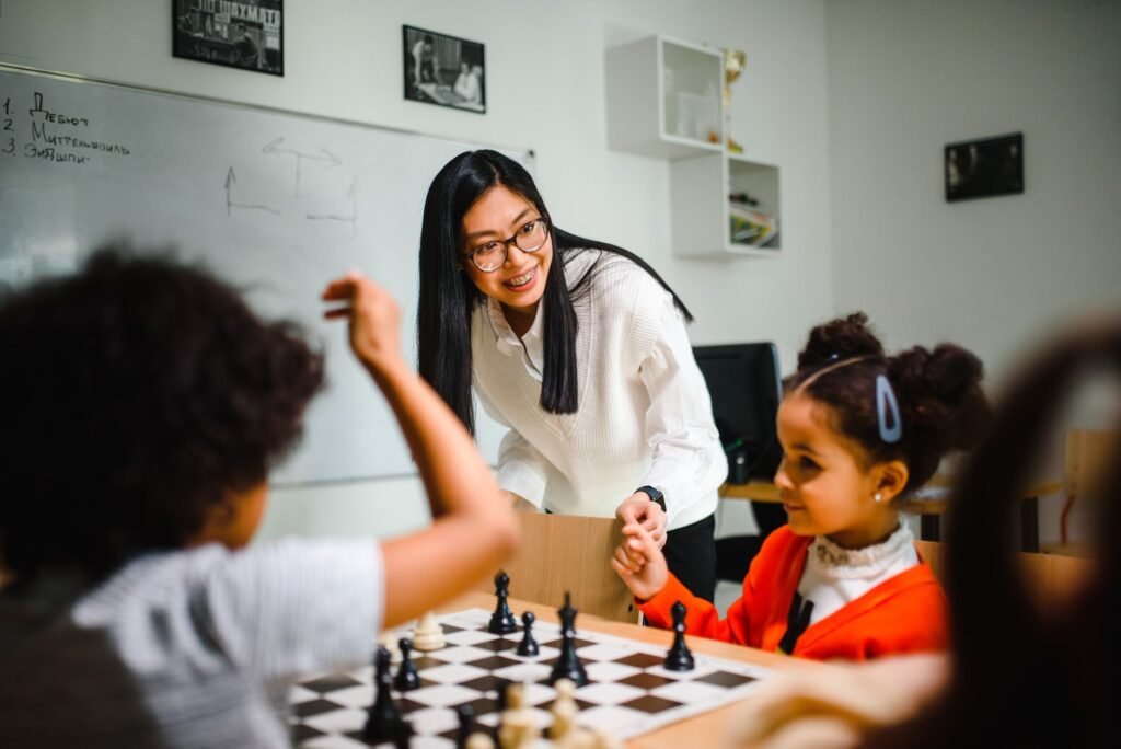 A chess coach guides young children at a board in a classroom setting, illustrating beginner chess instruction
